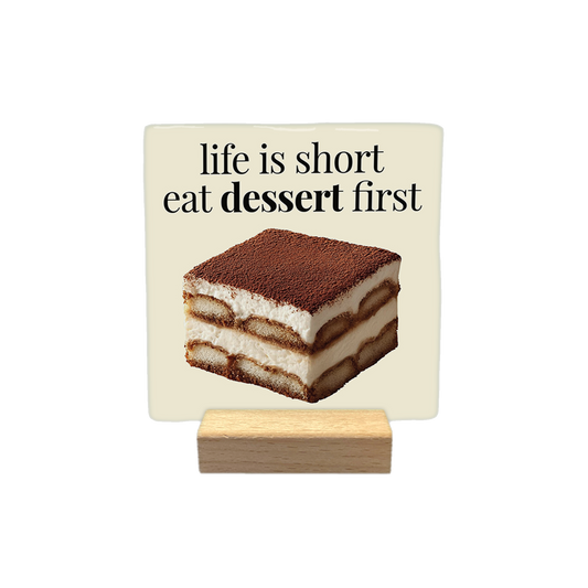 Life is short eat dessert first - Decoratie tegel - Formaat: - Op deze decoratietegel staat de tekst "life is short eat dessert first" in zwarte letters met een schreeflettertype. Daaronder is een realistische afbeelding van een vierkant stuk tiramisu geplaatst. Het ontwerp is gecentreerd op een crèmekleurige achtergrond. - Snelwenskaart