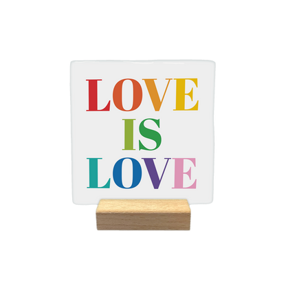 Love is Love - Decoratie tegel - Formaat: - Op deze decoratietegel staat de tekst "LOVE IS LOVE" in een groot schreeflettertype. Elk van de letters is uitgevoerd in een andere kleur van de regenboog, waaronder rood, oranje, geel, groen, blauw, paars en roze. - Snelwenskaart
