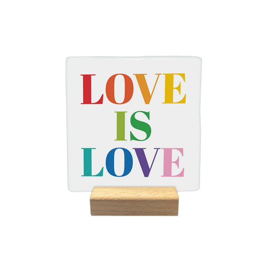 Love is Love - Decoratie tegel - Formaat: - Op deze decoratietegel staat de tekst "LOVE IS LOVE" in een groot schreeflettertype. Elk van de letters is uitgevoerd in een andere kleur van de regenboog, waaronder rood, oranje, geel, groen, blauw, paars en roze. - Snelwenskaart
