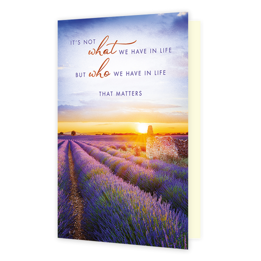 It's not what we have in life - Formaat: Standaard - Vorm: Staand - Vriendschapskaart - Op deze wenskaart staat een uitgestrekt, paars bloeiend lavendelveld met een stenen ruïne onder een warme zonsondergang. Boven het landschap staat de tekst "IT'S NOT what WE HAVE IN LIFE BUT who WE HAVE IN LIFE THAT MATTERS" in een combinatie van sierlijke oranje letters en strakke blauwe hoofdletters. De gouden lichtinval van de zon die achter de horizon verdwijnt, verlicht het veld en de lucht op een sfeervolle manier