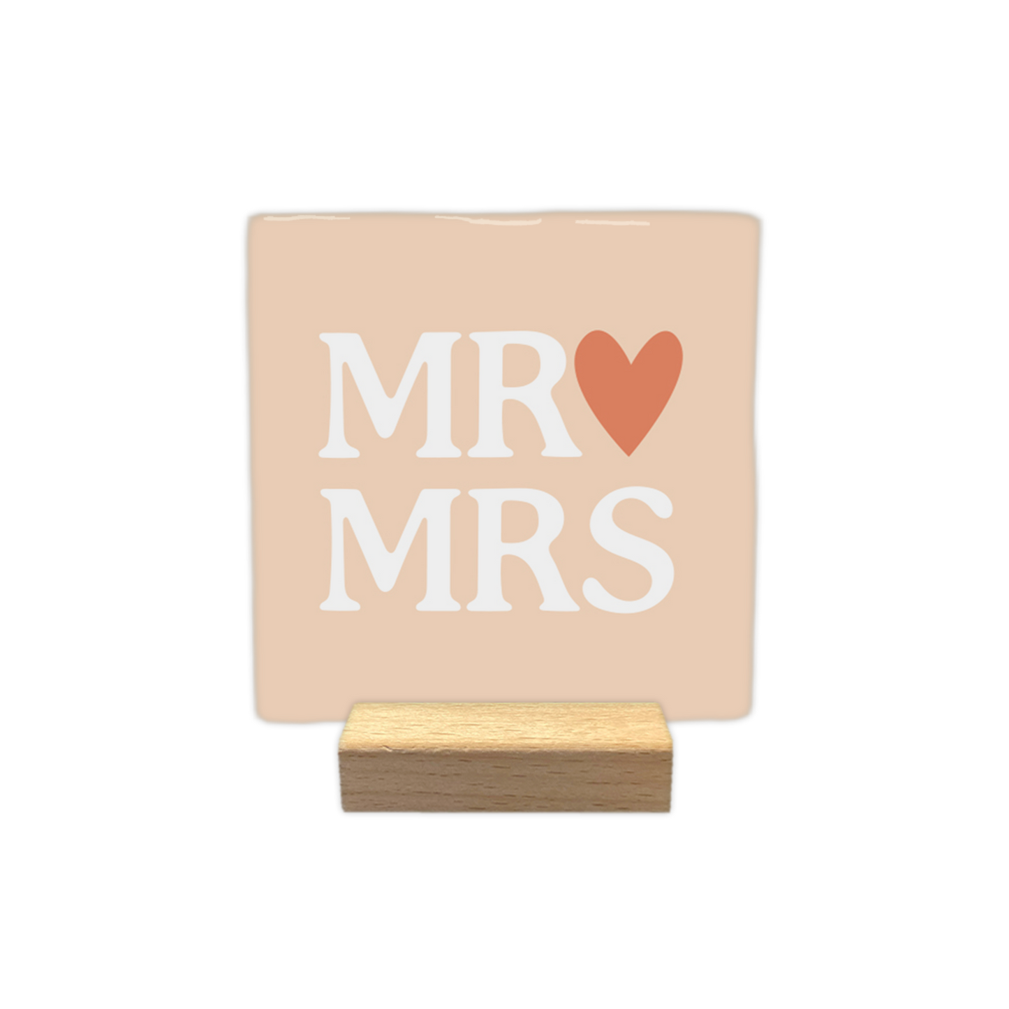 MR & MRS - Decoratie tegel - Formaat: - Op deze decoratietegel staat in het midden de Engelse tekst "MR MRS". De tegel zelf is zachtbeige van kleur, vierkant, en staat in een houten standaard. De tekst is verdeeld over twee regels en is geschreven in grote, witte hoofdletters. Direct achter het woord "MR" op de bovenste regel is een terracotta-oranje hartje afgebeeld. - Snelwenskaart