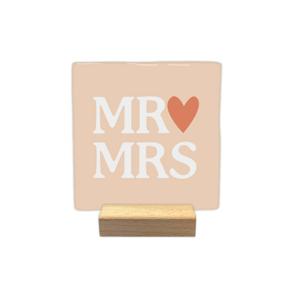 MR & MRS - Decoratie tegel - Formaat: - Op deze decoratietegel staat in het midden de Engelse tekst "MR MRS". De tegel zelf is zachtbeige van kleur, vierkant, en staat in een houten standaard. De tekst is verdeeld over twee regels en is geschreven in grote, witte hoofdletters. Direct achter het woord "MR" op de bovenste regel is een terracotta-oranje hartje afgebeeld. - Snelwenskaart