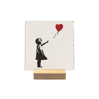 Meisje met ballon - Decoratie tegel - Formaat: - Op deze decoratie tegel staat een zwart silhouet van een jong meisje afgebeeld dat naar rechts kijkt. Ze staat op de grond en reikt met haar uitgestrekte rechterarm naar een rode hartvormige ballon. De ballon zweeft rechts boven haar aan een dun, golvend touwtje. - Snelwenskaart