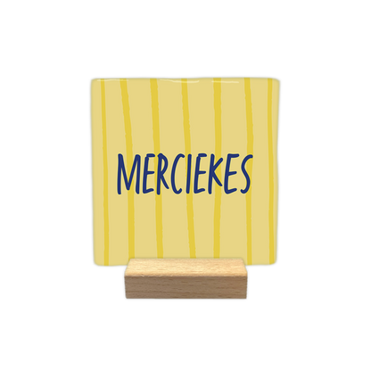 Merciekes - Decoratie tegel - Formaat: - Op deze decoratie tegel staat de tekst "merciekes" in blauwe, handgeschreven letters. De achtergrond van de tegel is lichtgeel en is voorzien van verticale, onregelmatige lijnen in een donkerdere kleur geel. - Snelwenskaart