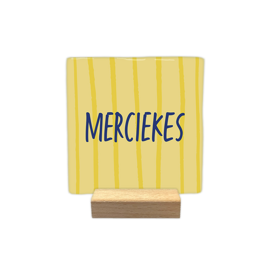 Merciekes - Decoratie tegel - Formaat: - Op deze decoratie tegel staat de tekst "merciekes" in blauwe, handgeschreven letters. De achtergrond van de tegel is lichtgeel en is voorzien van verticale, onregelmatige lijnen in een donkerdere kleur geel. - Snelwenskaart