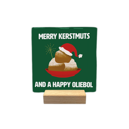 Merry kerstmuts - Decoratie tegel - Formaat: - Op deze decoratie tegel staat een groene achtergrond met een illustratie van een rode schaal met oliebollen erop. De bovenste oliebol draagt een rode kerstmuts met witte rand en er steekt een sterretje uit de schaal. Bovenaan en onderaan staat in witte letters de tekst merry kerstmuts and a happy oliebol. - Snelwenskaart