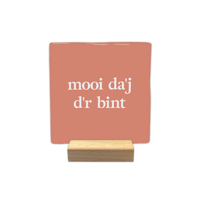 Mooi da'j d'r bint - Decoratie tegel - Formaat: - Op deze decoratie tegel staat de tekst "mooi da'j d'r bint" in witte letters verdeeld over twee regels. De achtergrond heeft een egale, zalmroze kleur. Het ontwerp is rustig en minimalistisch vormgegeven. - Snelwenskaart