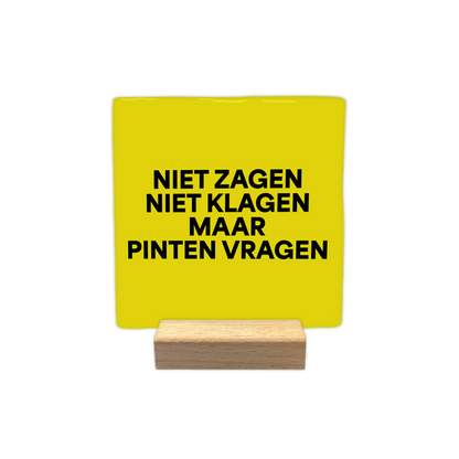 Niet zagen niet klagen maar pinten vragen - Decoratie tegel - Formaat: - Op deze decoratie tegel staat de tekst "niet zagen, niet klagen, maar pinten vragen" in vier regels afgebeeld. De tekst is geschreven in zwarte letters. De achtergrond van de tegel is geel. - Snelwenskaart