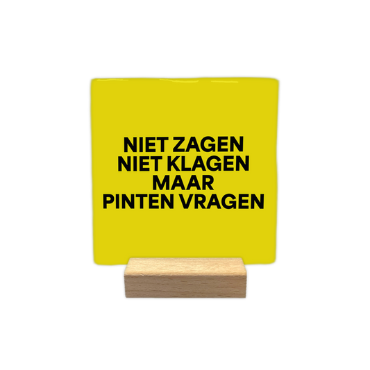 Niet zagen niet klagen maar pinten vragen - Decoratie tegel - Formaat: - Op deze decoratie tegel staat de tekst "niet zagen, niet klagen, maar pinten vragen" in vier regels afgebeeld. De tekst is geschreven in zwarte letters. De achtergrond van de tegel is geel. - Snelwenskaart