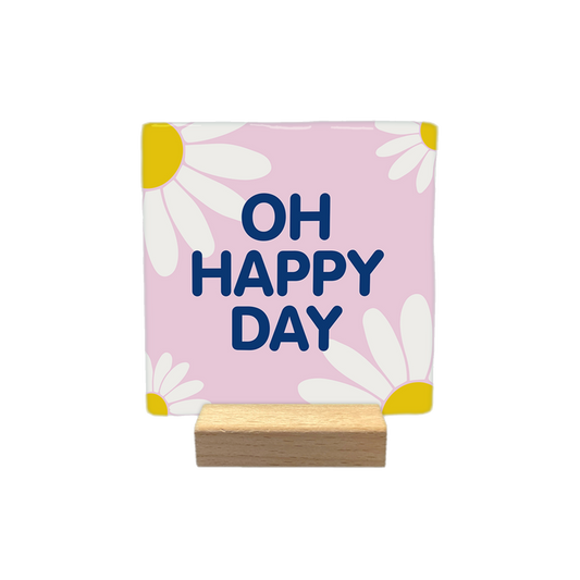 O happy day - Decoratie tegel - Formaat: - Op deze decoratietegel staat de tekst "OH HAPPY DAY" in donkerblauwe hoofdletters. De tekst is gecentreerd op een lichtroze, vierkante achtergrond die is versierd met gestileerde witte madeliefjes met gele harten in de hoeken. - Snelwenskaart