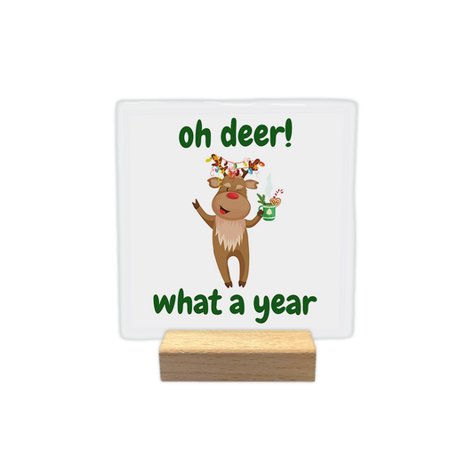 Oh deer! What a year - Decoratie tegel - Formaat: - Op deze decoratie tegel staat een lichte achtergrond met een illustratie van een rendier in het midden. Het rendier heeft kerstverlichting in zijn gewei en houdt een groene mok vast. Boven en onder het rendier staat in groene letters de tekst oh deer! what a year. - Snelwenskaart