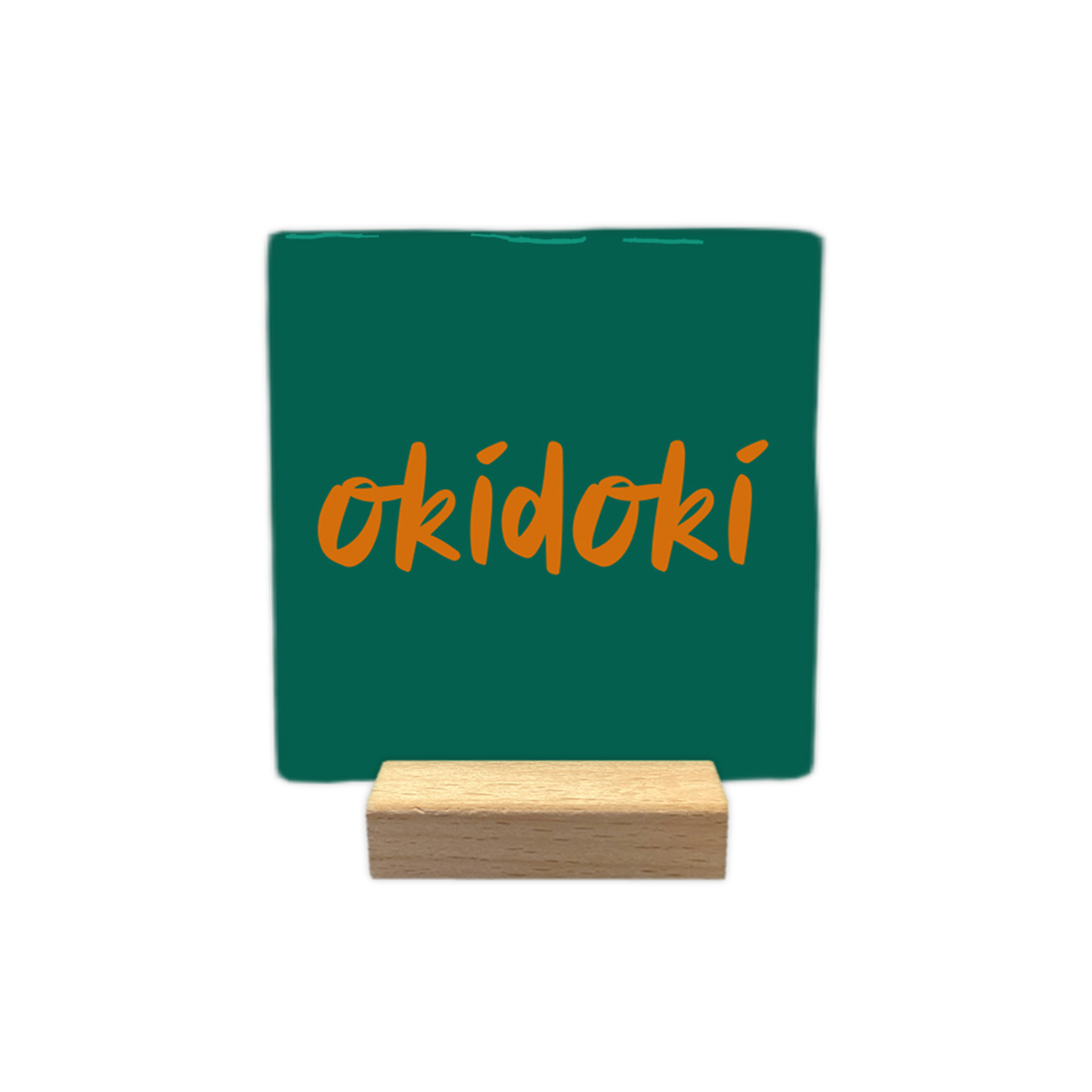 Okidoki - Decoratie tegel -  Op deze wenskaart is een vierkante, donkergroene decoratietegel afgebeeld tegen een egale, witte achtergrond. In het midden van de tegel staat het woord "okidoki" geschreven in een oranje, handgeschreven lettertype. - Snelwenskaart