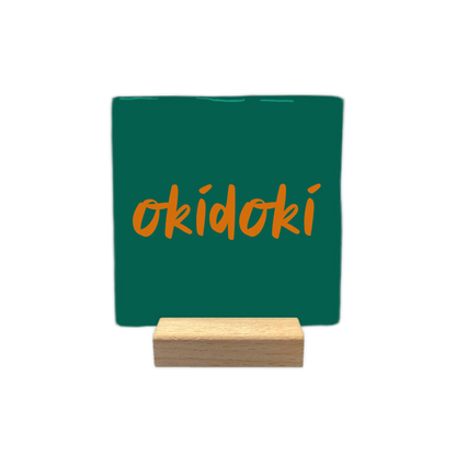 Okidoki - Decoratie tegel -  Op deze wenskaart is een vierkante, donkergroene decoratietegel afgebeeld tegen een egale, witte achtergrond. In het midden van de tegel staat het woord "okidoki" geschreven in een oranje, handgeschreven lettertype. - Snelwenskaart
