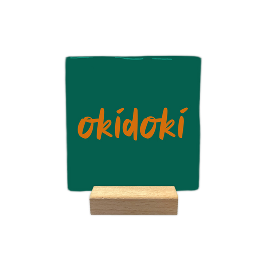 Okidoki - Decoratie tegel -  Op deze wenskaart is een vierkante, donkergroene decoratietegel afgebeeld tegen een egale, witte achtergrond. In het midden van de tegel staat het woord "okidoki" geschreven in een oranje, handgeschreven lettertype. - Snelwenskaart