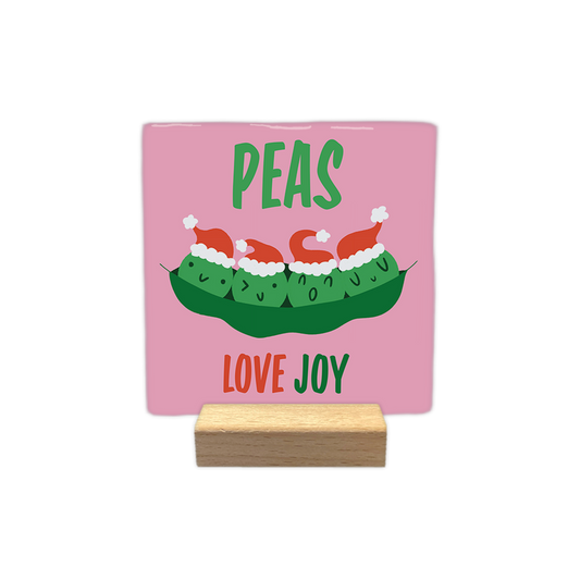 Peas love joy - Decoratie tegel - Formaat: - Op deze decoratie tegel staat een roze achtergrond met daarin een groene peul met vier erwten. Elke erwt draagt een klein rood kerstmutsje met een witte rand en pompon en heeft een eigen gezichtje. Boven de peul staat in groene letters de tekst peas. Onder de peul staat in rode en groene letters de tekst love joy. - Snelwenskaart