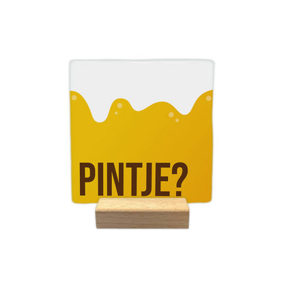 Pintje? - Decoratie tegel - Formaat: - Op deze decoratie tegel is onderaan de tekst "pintje?" in donkerbruine letters afgebeeld. De rest van de tegel is vormgegeven als een glas bier, met een gele vloeistof die aan de bovenkant golft en kleine luchtbelletjes bevat. De achtergrond van het bovenste gedeelte is wit. - Snelwenskaart