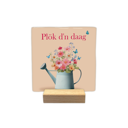 Plök d'n daag - Decoratie tegel - Formaat: - Op deze decoratie tegel staat de tekst "Plök d'n daag" in roze letters. Onder de tekst staat een lichtblauwe gieter die gevuld is met een kleurrijk boeket van diverse bloemen. Het ontwerp wordt opgefleurd door verschillende kleine, blauwe vlinders die rond de bloemen vliegen. De achtergrond heeft een zachte, beige kleur. - Snelwenskaart