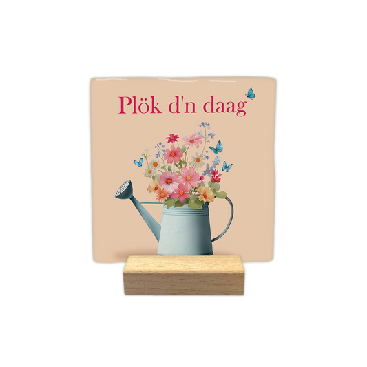 Plök d'n daag - Decoratie tegel - Formaat: - Op deze decoratie tegel staat de tekst "Plök d'n daag" in roze letters. Onder de tekst staat een lichtblauwe gieter die gevuld is met een kleurrijk boeket van diverse bloemen. Het ontwerp wordt opgefleurd door verschillende kleine, blauwe vlinders die rond de bloemen vliegen. De achtergrond heeft een zachte, beige kleur. - Snelwenskaart