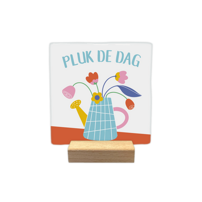 Pluk de dag - Decoratie tegel - Formaat:  10 x 10 - Op deze decoratietegel staat bovenaan in een lichte boog de Nederlandse tekst "PLUK DE DAG". De tegel zelf is wit en vierkant, en staat in een houten standaard. In het midden is een kleurrijke illustratie te zien van een lichtblauwe, geblokte gieter die als vaas dient voor verschillende getekende bloemen in de kleuren roze, rood en geel. De gieter heeft een gele tuit en een roze handvat, en staat op een schuine, terracotta-oranje ondergrond.- Snelwenskaart