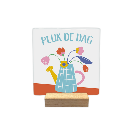 Pluk de dag - Decoratie tegel - Formaat:  10 x 10 - Op deze decoratietegel staat bovenaan in een lichte boog de Nederlandse tekst "PLUK DE DAG". De tegel zelf is wit en vierkant, en staat in een houten standaard. In het midden is een kleurrijke illustratie te zien van een lichtblauwe, geblokte gieter die als vaas dient voor verschillende getekende bloemen in de kleuren roze, rood en geel. De gieter heeft een gele tuit en een roze handvat, en staat op een schuine, terracotta-oranje ondergrond.- Snelwenskaart