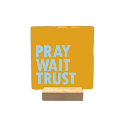 Pray wait trust - Decoratie tegel - Formaat: - Op deze decoratie tegel staat de tekst "pray", "wait" en "trust" onder elkaar in lichtblauwe letters. De woorden zijn links uitgelijnd en uitgevoerd in een strak, vet lettertype. De achtergrond van de tegel is een egale, heldergele kleur. - Snelwenskaart