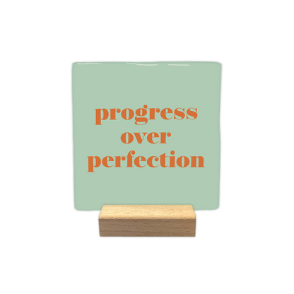 Progress over perfection - Decoratie tegel - Formaat: - Op deze decoratietegel staat de tekst "progress over perfection" in kleine letters met een schreeflettertype. De tekst is uitgevoerd in een oranje kleur en is gecentreerd op een mintgroene achtergrond. - Snelwenskaart
