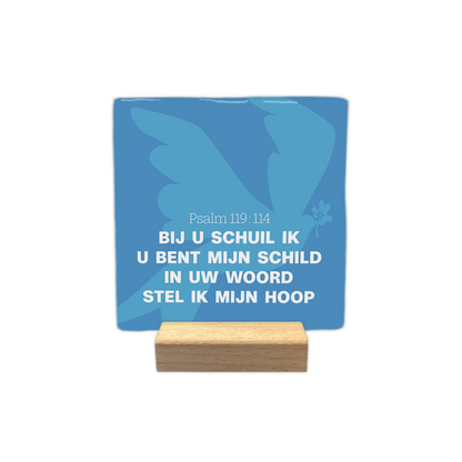 Psalm 119:114 - Decoratie tegel - Formaat: - Op deze decoratie tegel staat een lichtblauwe afbeelding van een vogel met gespreide vleugels tegen een donkerblauwe achtergrond, met een takje met bladeren in de snavel. In het midden is de tekst Psalm 119:114 in witte letters weergegeven. Daaronder staat de tekst "Bij u schuil ik", "U bent mijn schild", "In uw woord" en "Stel ik mijn hoop" in witte hoofdletters en kleine letters. Alle tekst op de tegel is in het wit uitgevoerd. - Snelwenskaart