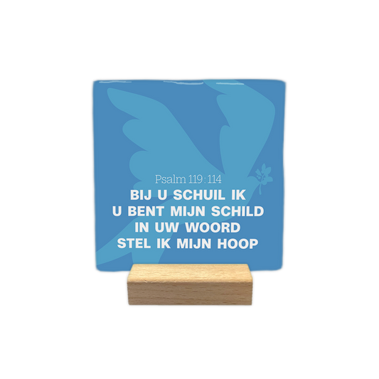 Psalm 119:114 - Decoratie tegel - Formaat: - Op deze decoratie tegel staat een lichtblauwe afbeelding van een vogel met gespreide vleugels tegen een donkerblauwe achtergrond, met een takje met bladeren in de snavel. In het midden is de tekst Psalm 119:114 in witte letters weergegeven. Daaronder staat de tekst "Bij u schuil ik", "U bent mijn schild", "In uw woord" en "Stel ik mijn hoop" in witte hoofdletters en kleine letters. Alle tekst op de tegel is in het wit uitgevoerd. - Snelwenskaart
