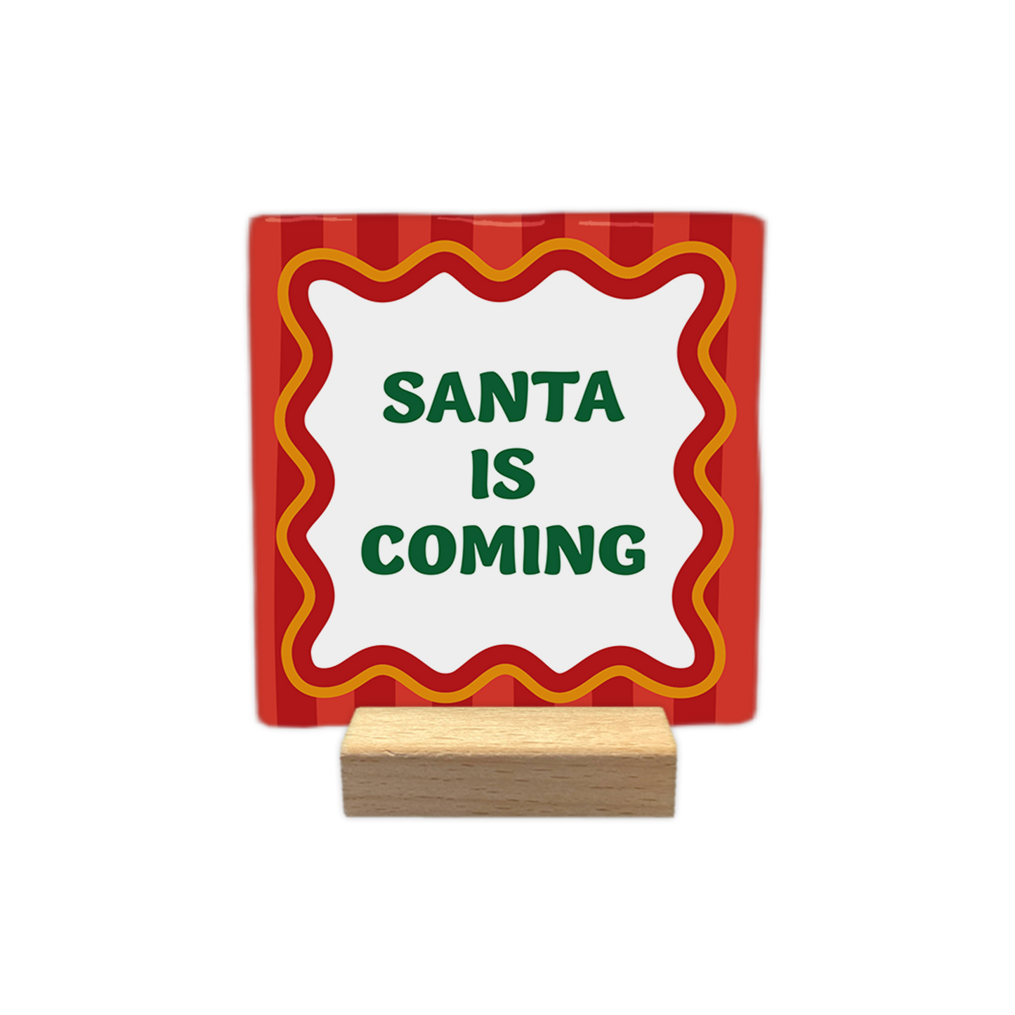 Santa is coming - Decoratie tegel - Op deze decoratietegel staat centraal de tekst "SANTA IS COMING" in groene letters. De tekst is omgeven door een wit kader met een golvende rand, dat op zijn beurt weer een rood-oranje omlijning heeft. De achtergrond van de tegel is versierd met een patroon van verticale rode strepen.- Snelwenskaart