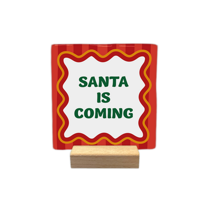 Santa is coming - Decoratie tegel - Op deze decoratietegel staat centraal de tekst "SANTA IS COMING" in groene letters. De tekst is omgeven door een wit kader met een golvende rand, dat op zijn beurt weer een rood-oranje omlijning heeft. De achtergrond van de tegel is versierd met een patroon van verticale rode strepen.- Snelwenskaart