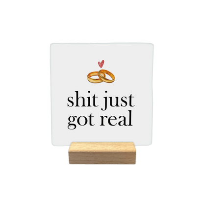 Shit just got real - Decoratie tegel - Formaat: - Op deze decoratietegel staat de tekst "shit just got real" in zwarte letters met een klassiek schreeflettertype. Boven de tekst zijn twee in elkaar grijpende trouwringen afgebeeld, met daarboven een klein, handgetekend rood hartje. - Snelwenskaart