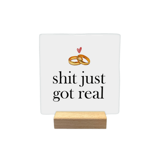 Shit just got real - Decoratie tegel - Formaat: - Op deze decoratietegel staat de tekst "shit just got real" in zwarte letters met een klassiek schreeflettertype. Boven de tekst zijn twee in elkaar grijpende trouwringen afgebeeld, met daarboven een klein, handgetekend rood hartje. - Snelwenskaart