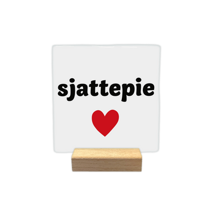 Sjattepie - Decoratie tegel - Formaat: - Op deze decoratie tegel staat de tekst "sjattepie" in zwarte letters tegen een witte achtergrond. Onder de tekst is een rood hart afgebeeld. Het ontwerp is eenvoudig en strak vormgegeven. - Snelwenskaart