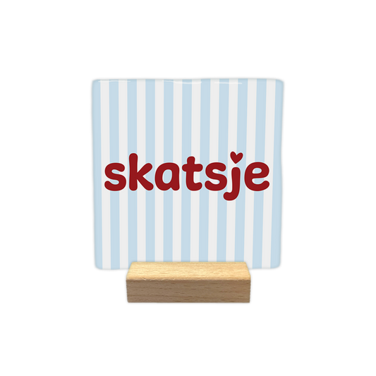 Skatsje - Decoratie tegel - Formaat: - Op deze decoratie tegel staat de tekst "skatsje" in rode letters centraal afgebeeld. Boven de letter j bevindt zich een klein hartje. De achtergrond van de tegel is voorzien van verticaal gestreepte blauwe en witte banen. - Snelwenskaart