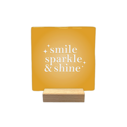 Smile sparkle & shine - Decoratie tegel - Formaat: - Op deze decoratietegel staat de tekst "smile sparkle & shine" in witte letters met een schreeflettertype. De tekst is gecentreerd op een goudgele, vierkante achtergrond en wordt aan weerszijden vergezeld door kleine, gestileerde sterretjes. - Snelwenskaart