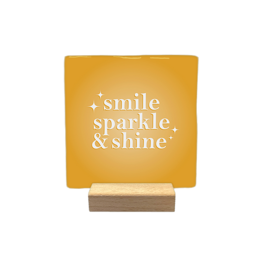 Smile sparkle & shine - Decoratie tegel - Formaat: - Op deze decoratietegel staat de tekst "smile sparkle & shine" in witte letters met een schreeflettertype. De tekst is gecentreerd op een goudgele, vierkante achtergrond en wordt aan weerszijden vergezeld door kleine, gestileerde sterretjes. - Snelwenskaart