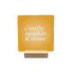 Smile sparkle & shine - Decoratie tegel - Formaat: - Op deze decoratietegel staat de tekst "smile sparkle & shine" in witte letters met een schreeflettertype. De tekst is gecentreerd op een goudgele, vierkante achtergrond en wordt aan weerszijden vergezeld door kleine, gestileerde sterretjes. - Snelwenskaart