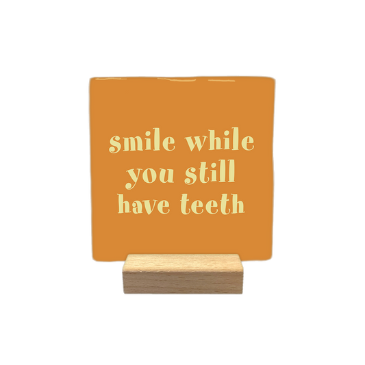 Smile while you still have teeth - Decoratie tegel - Formaat: - Op deze decoratietegel staat de tekst "smile while you still have teeth" in lichtgele letters. De tekst is gecentreerd op een oranje achtergrond. - Snelwenskaart