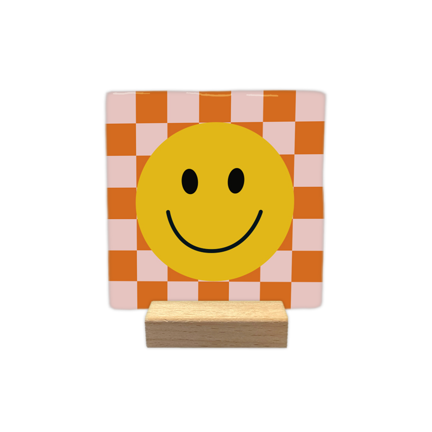 Smiley - Decoratie tegel - Formaat: - Op deze decoratietegel staat centraal een grote, gele smiley afgebeeld met zwarte ogen en een glimlachende mond. De achtergrond van de illustratie bestaat uit een schaakbordpatroon van afwisselend oranje en lichtroze vierkanten. - Snelwenskaart