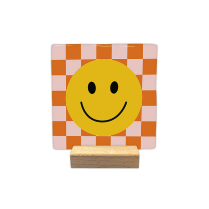 Smiley - Decoratie tegel - Formaat: - Op deze decoratietegel staat centraal een grote, gele smiley afgebeeld met zwarte ogen en een glimlachende mond. De achtergrond van de illustratie bestaat uit een schaakbordpatroon van afwisselend oranje en lichtroze vierkanten. - Snelwenskaart