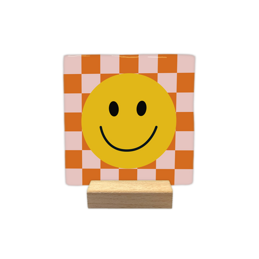 Smiley - Decoratie tegel - Formaat: - Op deze decoratietegel staat centraal een grote, gele smiley afgebeeld met zwarte ogen en een glimlachende mond. De achtergrond van de illustratie bestaat uit een schaakbordpatroon van afwisselend oranje en lichtroze vierkanten. - Snelwenskaart