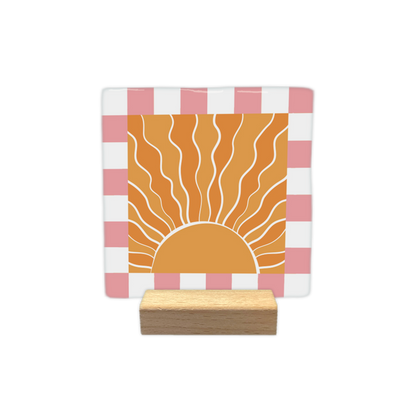 Sunshine - Decoratie tegel - Formaat: - Op deze decoratietegel staat een gestileerde, opkomende zon centraal afgebeeld in een oranje vierkant. De zon is getekend met golvende, witte stralen die naar boven uitstralen vanuit een halve cirkel aan de onderzijde. Het centrale oranje vierkant is omgeven door een rand met een schaakbordpatroon van afwisselende witte en roze vierkanten. - Snelwenskaart