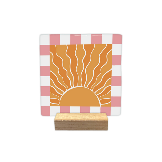 Sunshine - Decoratie tegel - Formaat: - Op deze decoratietegel staat een gestileerde, opkomende zon centraal afgebeeld in een oranje vierkant. De zon is getekend met golvende, witte stralen die naar boven uitstralen vanuit een halve cirkel aan de onderzijde. Het centrale oranje vierkant is omgeven door een rand met een schaakbordpatroon van afwisselende witte en roze vierkanten. - Snelwenskaart