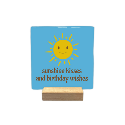Sunshine kisses and birthday wishes - Decoratie tegel - Formaat: - Op deze decoratie tegel staat in het bruin de tekst 'Sunshine kisses and birthday wishes'. Boven de tekst staat een gele zon met gezicht. De achtergrond van de decoratie tegel is blauw. - Snelwenskaart