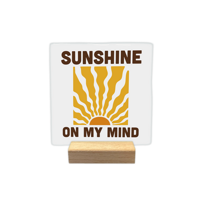 Sunshine on my mind - Decoratie tegel - Formaat: - Op deze decoratie tegel staat een gestileerde afbeelding van een opkomende zon met golvende zonnestralen die naar boven uitstralen. Zowel boven als onder deze illustratie staat de tekst "SUNSHINE ON MY MIND" in grote, donkerbruine hoofdletters. De zon en de stralen zijn weergegeven in warme tinten oranje en geel tegen een witte achtergrond. - Snelwenskaart