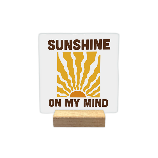 Sunshine on my mind - Decoratie tegel - Formaat: - Op deze decoratie tegel staat een gestileerde afbeelding van een opkomende zon met golvende zonnestralen die naar boven uitstralen. Zowel boven als onder deze illustratie staat de tekst "SUNSHINE ON MY MIND" in grote, donkerbruine hoofdletters. De zon en de stralen zijn weergegeven in warme tinten oranje en geel tegen een witte achtergrond. - Snelwenskaart