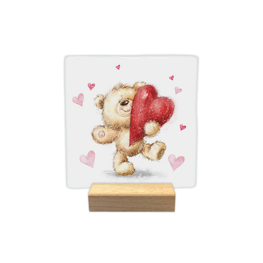 Decoratietegel – 10 x 10 cm - Op deze decoratietegel staat een illustratie van een pluizig, lichtbruin teddybeertje dat een groot, glinsterend rood hart vasthoudt. Het beertje is omringd door diverse kleine, roze hartjes tegen een witte achtergrond. Het ontwerp heeft een zachte, aquarelachtige stijl.