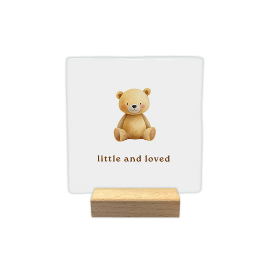 Decoratietegel – 10 x 10 cm - Op deze decoratietegel staat onderaan de Engelse tekst "little and loved". De tegel zelf is wit en vierkant, en staat in een houten standaard. Boven de tekst is een schattige illustratie te zien van een zittende teddybeer in zachte bruintinten, compleet met kleine blosjes op de wangen. De tekst is op één regel geschreven in een strak, bruin lettertype.