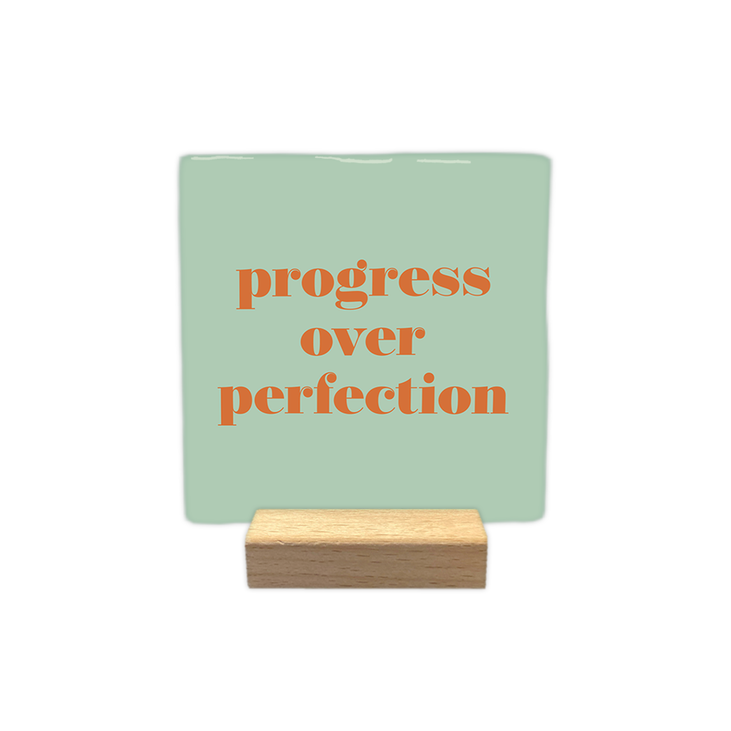 Decoratietegel – 10 x 10 cm - Op deze decoratietegel staat de tekst "progress over perfection" in kleine letters met een schreeflettertype. De tekst is uitgevoerd in een oranje kleur en is gecentreerd op een mintgroene achtergrond. 