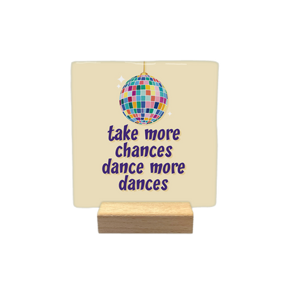 Decoratietegel – 10 x 10 cm - Op deze decoratietegel staat een afbeelding van een kleurrijke discobal boven de tekst "take more chances dance more dances" in paarse letters. De tekst en de discobal zijn gecentreerd op een lichtbeige achtergrond.